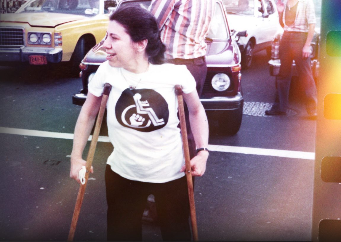 Photo extraite de "Crip Camp" : Sur la chaussée, une femme soutenue par deux cannes. Son tee-shirt porte le pictogramme "personne à mobilité réduite" avec un poing fermé au centre de la roue. Derrière elle, des passants et des voitures à l'arrêt.