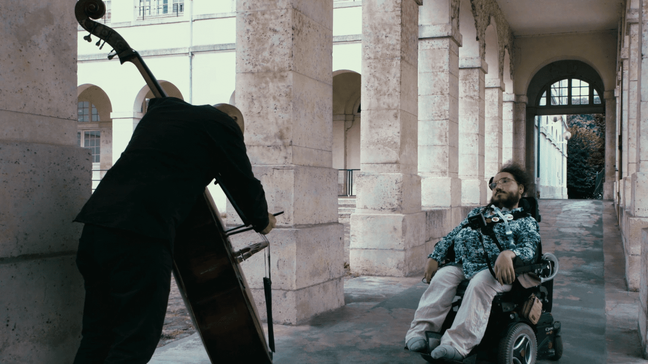 Photo du film "Fils de Garches" : Rémi Gendarme assis dans son fauteuil roulant, au bas d'un large plan incliné. Face à lui, un homme vêtu de noir, le dos courbé sur une contrebasse.