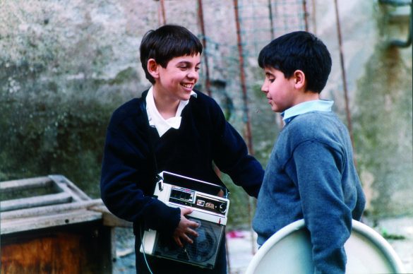 Photo extraite du film : Deux jeunes garçons se font face. Celui de gauche arbore un grand sourire. Il porte un magnétophone en bandoulière sur lequel il pose une main. De profil, celui de droite a la paupière baissée.