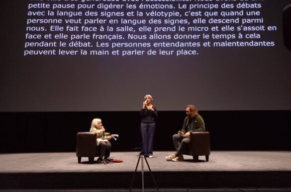 Photo : Au pied de l'écran de cinéma sur lequel figure la transcription des échanges, Diane Maroger et Nicolas Becker, assis sur des fauteuils. Entre eux, l'interprète en LSF.