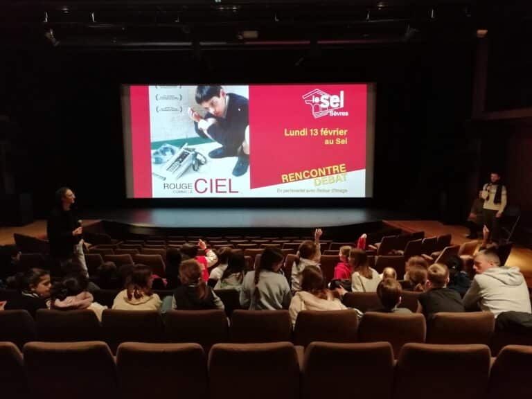 Photo : L’affiche du film "Rouge comme le ciel" est projetée sur l’écran de la salle. Des mains dans le public sont levées. Sur la scène, à droite de l’écran, l’intervenant Alexis Robin, micro à la main.