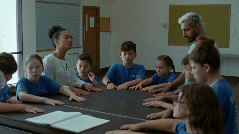 Photo extraite du film : autour d'une table grise, Ruben et une professeure sont assis parmi des enfants vêtus de bleu. Tous ont les mains posées sur la table. Certains ferment les yeux.