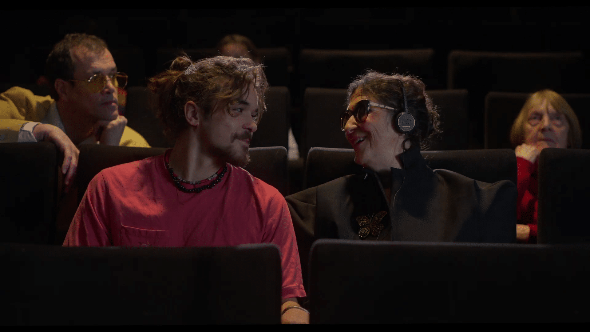 Image extraite du film "Au-delà du regard" de Andres Hernandez Machado : Dans une salle de cinéma, un homme et une femme se regardent en souriant. Casque sur les oreilles, la femme porte des lunettes noires.