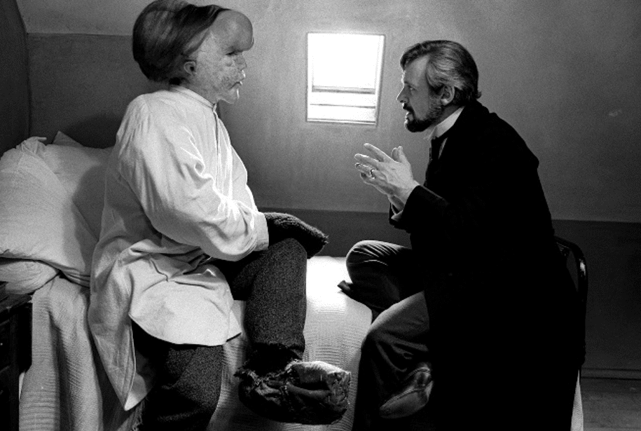 Photo en noir et blanc du film Elephant Man : deux hommes sont assis face à face et se regardent. Celui de gauche, le visage déformé, porte une chemise blanche. Il est installé sur le bord d'un lit. Celui de droite, vêtu d'un costume sombre, est assis sur une chaise. Derrière, entre les deux hommes, une fenêtre rectangulaire filtre la lumière du jour.