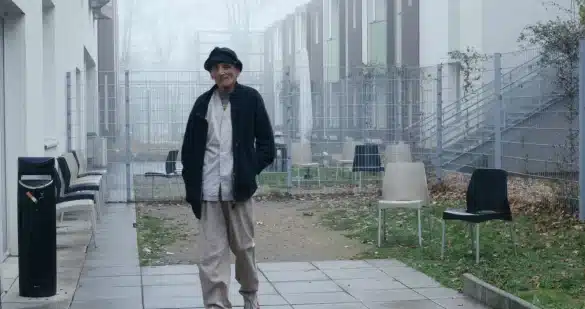 Photo extraite du film 12 jours : Dans une cour délimitée par un grillage, un homme marche. Il porte un chapeau et une veste sombres, une chemise blanche et un pantalon clair. Dans un coin de la cour, une chaise blanche et une chaise noire sont disposées près de la clôture. Au second plan, le paysage est embrumé.