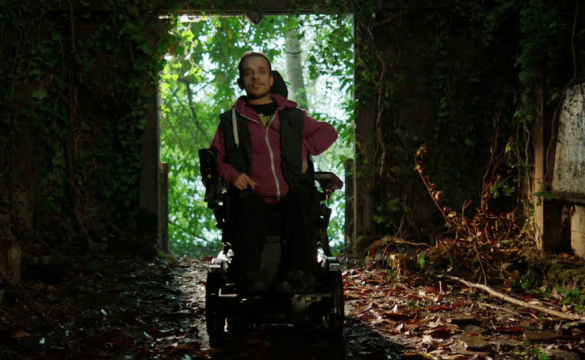 Photo extraite du film Je d'un accident ou d'amour : dans un bâtiment abandonné et sombre, envahit par la nature, un homme en fauteuil roulant se tient dans l'encadrement d'une porte qui laisse entrer la lumière du jour.