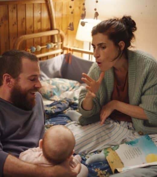 Photo extraite de Sorda : dans une chambre d'enfant, une homme et une femme sont assis sur un lit. L'homme tient un bébé dans son bras. La femme signe d'une main. Un livre illustré est ouvert devant eux.