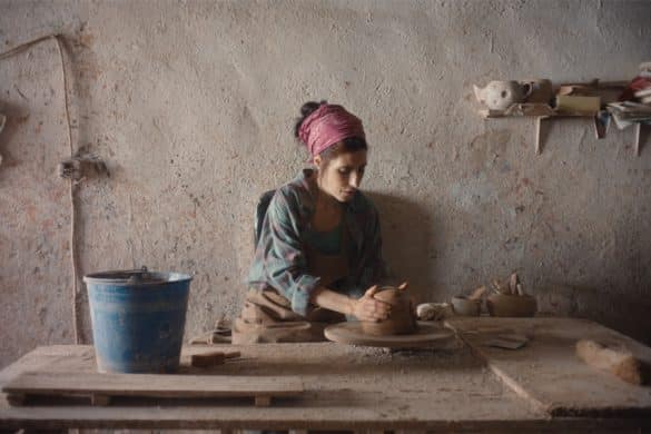 Photo extraite de Sorda : Dans un atelier au mur beige, un foulard rose dans les cheveux, une femme est assise derrière une table en bois. Ses mains sont placées sur un morceau d'argile. Un seau bleu est posé près d'elle.