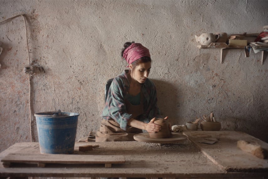 Photo extraite de Sorda : Dans un atelier au mur beige, un foulard rose dans les cheveux, une femme est assise derrière une table en bois. Ses mains sont placées sur un morceau d'argile. Un seau bleu est posé près d'elle.