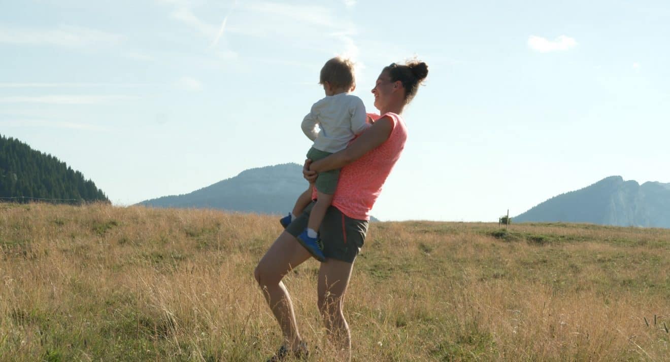 Photo extraite de Elle entend pas la moto : Dans un champs aux herbes jaunies, et sur fond de ciel clair, une femme au tee-shirt rose porte un enfant dans ses bras.