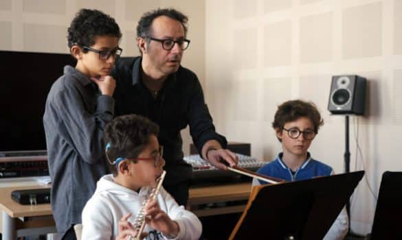 Photo extraite du film La musique sourde : un homme tient un archet à la main, il le pointe vers un pupitre. Il est entouré de trois garçons. L'un d'eux porte une flûte traversière à ses lèvres.