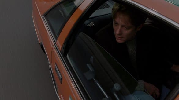 Photo extraite du film Crash : un homme assis à l'arrière d'une voiture orange regarde par l'encadrure de la fenêtre ouverte. La voiture est sur une voie en bitume.