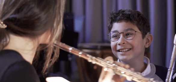 Photo extraite du film la musique sourde : Un garçon brun portant des lunettes arbore un grand sourire. Au premier plan, de dos, une femme joue de la flûte traversière.