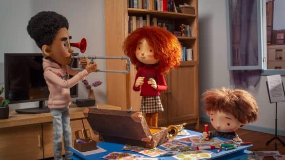 Photo extraite du film Lola et la piano à bruits (film d'animation en stop motion) : trois enfants dans un salon. Un garçon à la peu brune porte un trombone à ses lèvres. Une fille rousse se tient debout à ses côtés. Assis par terre, à une table basse, un garçon au teint blanc, plus jeune que les deux autres, tient un crayon au-dessus d'une feuille colorée.