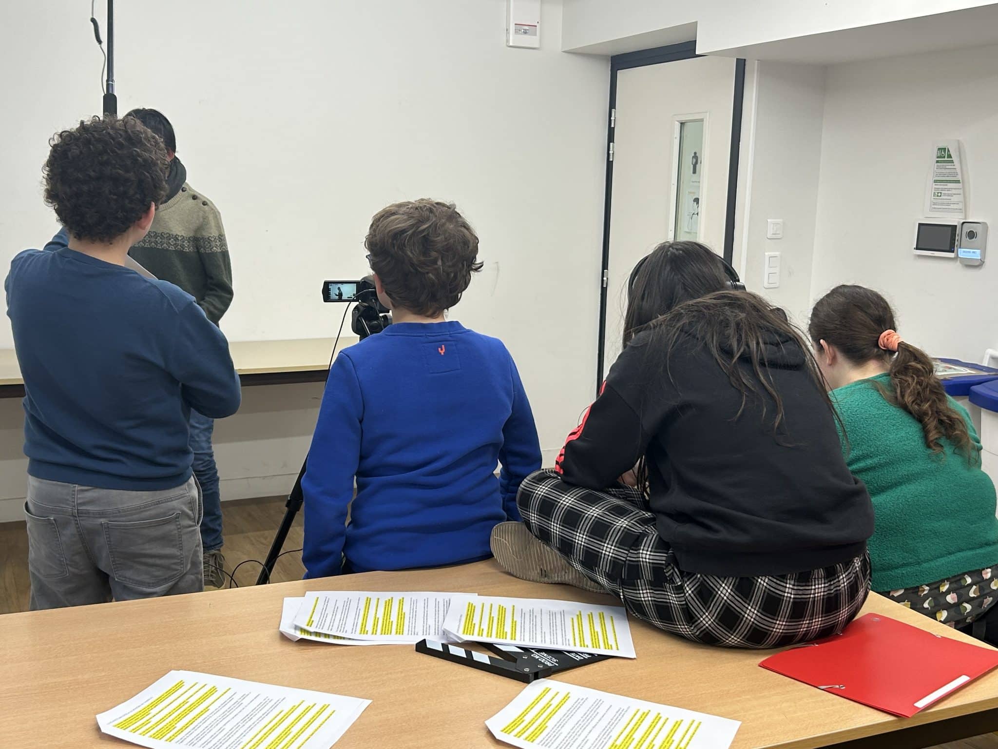 Photo de l'atelier : Quatre élèves de dos ; deux sont debout derrière une caméra, une autre assise sur une table, et la dernière sur une chaise à ses côtés. Des feuilles de papier remplies de texte sont disposées sur la table.