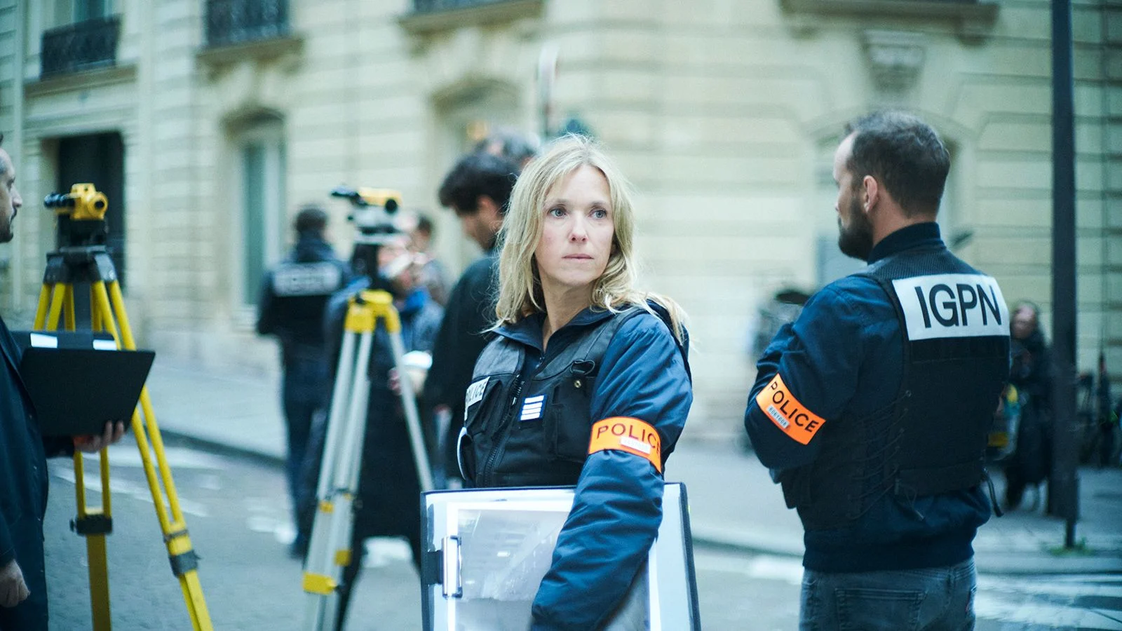 Photo de Dossier 137 : Dans une rue, une femme blonde avec une veste noire et un brassard orange. Elle porte un dossier sous le bras. Un homme se tient de dos à ses côtés. Les lettres IGPN sont inscrites sur sa veste noire, et le mot Police sur les brassards des deux personnes.