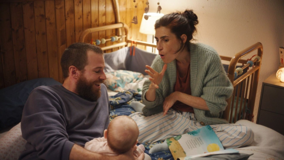 Photo extraite de Sorda : dans une chambre d'enfant, une homme et une femme sont assis sur un lit. L'homme tient un bébé dans son bras. La femme signe d'une main. Un livre illustré est ouvert devant eux.
