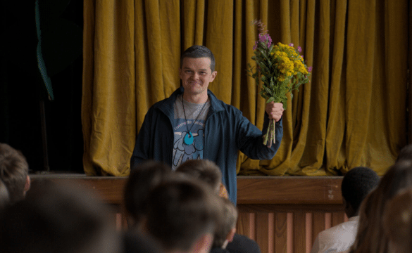 Photo extraite du film Plus fort que moi : Debout au pied d'un rideau jaune, un homme aux cheveux courts légèrement grisés tient un bouquet de fleurs jaunes et violettes dans sa main levée à hauteur d'épaule. Au premier plan, de dos, les têtes des spectateurs.