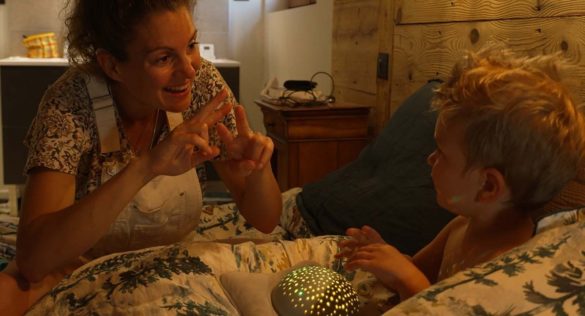 Photo extraite du film Elle entend pas la moto : Assis en tailleur sur la couette d'un lit, une femme et un enfant blonds se regardent. Souriante, la femme s'exprime en langue des signes.