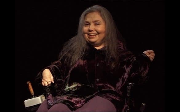 Photo de Cheryl Marie Wade : Un plateau sombre. Une femme d'une cinquantaine d'année dans un grand fauteuil électrique vintage. Cadré de face jusqu'au cuisse, elle porte une chemise de velours foncée et une longue jupe violette.