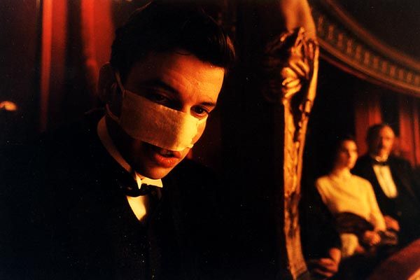 Photo extrait du film La chambre des officiers : Dans une loge de théâtre, un homme porte un bandage qui lui couvre le visage au niveau du nez. Il est vêtu d'un costume noir. Une lumière chaude orangée éclaire son visage. Dans la loge voisine, en arrière-plan flou, se tiennent une femme et un homme en tenue de soirée.