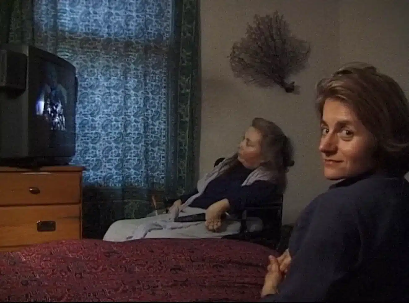Photo : De dos, dans le coin droit de l'image, Diane Maroger tourne le visage vers la caméra. En arrière-plan, Cheryl Marie Wade est assise dans son fauteuil, et regarde un écran de télévision.