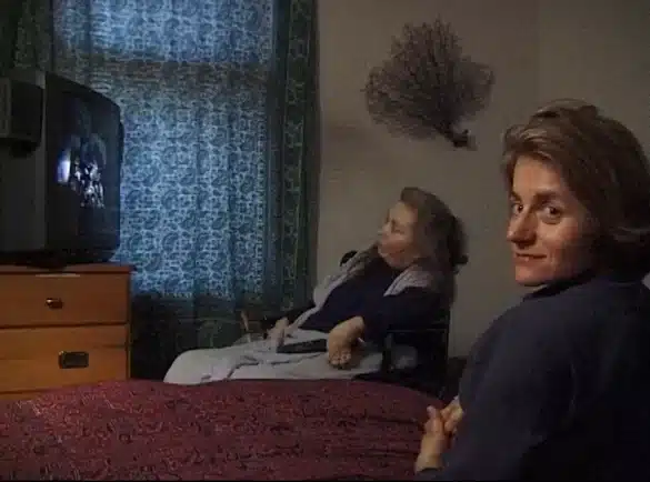 Photo : De dos, dans le coin droit de l'image, Diane Maroger tourne le visage vers la caméra. En arrière-plan, Cheryl Marie Wade est assise dans son fauteuil, et regarde un écran de télévision.