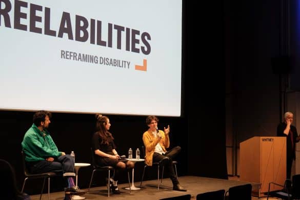 Photo du festival : sur scène devant un écran blanc sur lequel est inscrit Reelabilities, Reframing disability, les trois intervenants sont assis. Parmi eux, en veste jaune et pantalon noir, un micro à la main : le réalisateur Andrés Hernandez Machado.