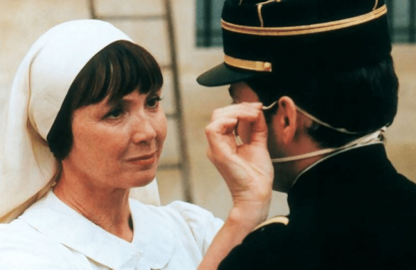 Photo extraite du film la chambre des officiers : le buste d'une femme brune en habit blanc d'infirmière, et celui d'un homme brun, de dos, portant un uniforme militaire noir et un képi avec des lignes dorées. La femme tend la main droite pour ajuster le masque que l'homme porte sur son visage.
