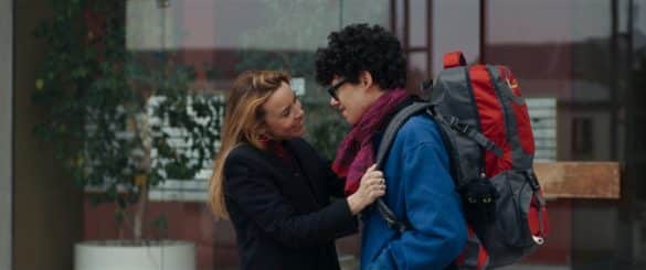 Photo extraite du film Ulysse : au centre-gauche de l'image une femme blonde réajuste le sac à dos d'un adolescent brun, son fils, qui se tient devant elle.