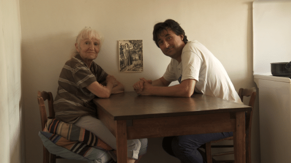 Photo extraite du film Eredita : deux personnes sont assises l'une en face de l'autre à une table et sont tournées vers nous. A gauche, Muguette, à droite Jean-Luc Cesco.