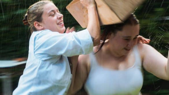 Photo extraite du film Au bord du monde : deux jeunes femmes s'amusent sous la pluie. La jeune femme à gauche de l'image semble vouloir placer une boîte en carton sur la tête de la jeune femme à droite de l'image.