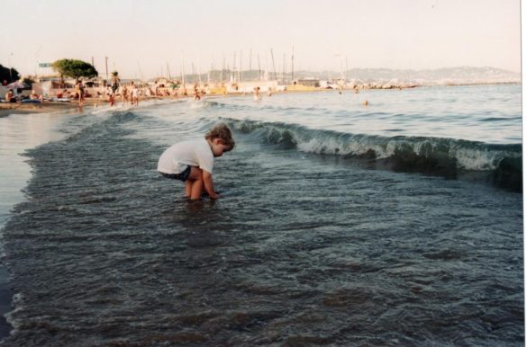 Image de Comme une pieuvre : une enfant est accroupie à gauche de l'image, les pieds dans la mer, derrière elle un port.